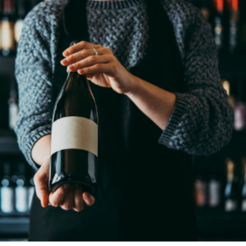 A person wearing a grey sweater under a black apron holds glass wine bottle in front of a shelf full of wine bottles