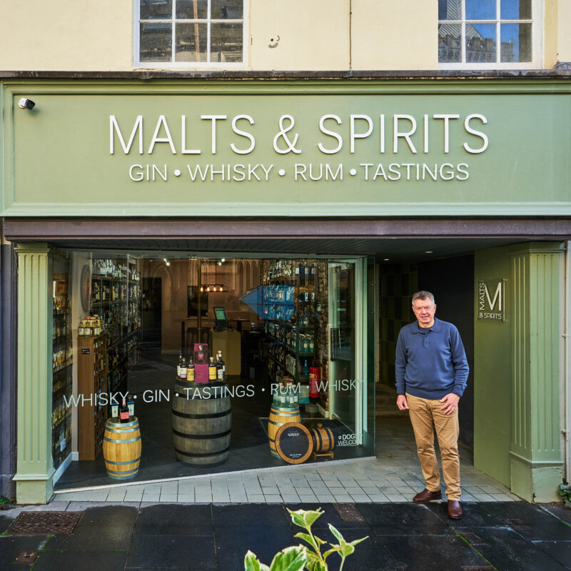 A man wearing a grey jumper stands outside a shop painted light green with a large window displaying bottles of alcohol