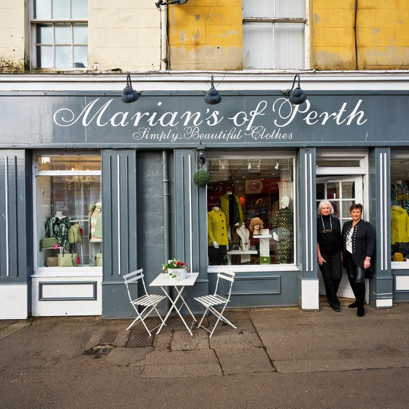 Two smiling women wearing black clothes stand outside a grey shop with large windows displaying an array of colourful clothing