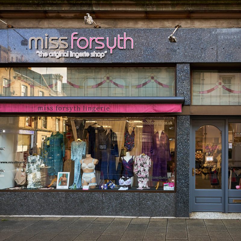 A granite shop front with pink signs and a decorated window above a large window displaying women's lingerie.