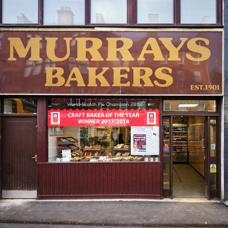A large brown sign with a yellow font sits above a bakery window displaying an assortment of baked goods
