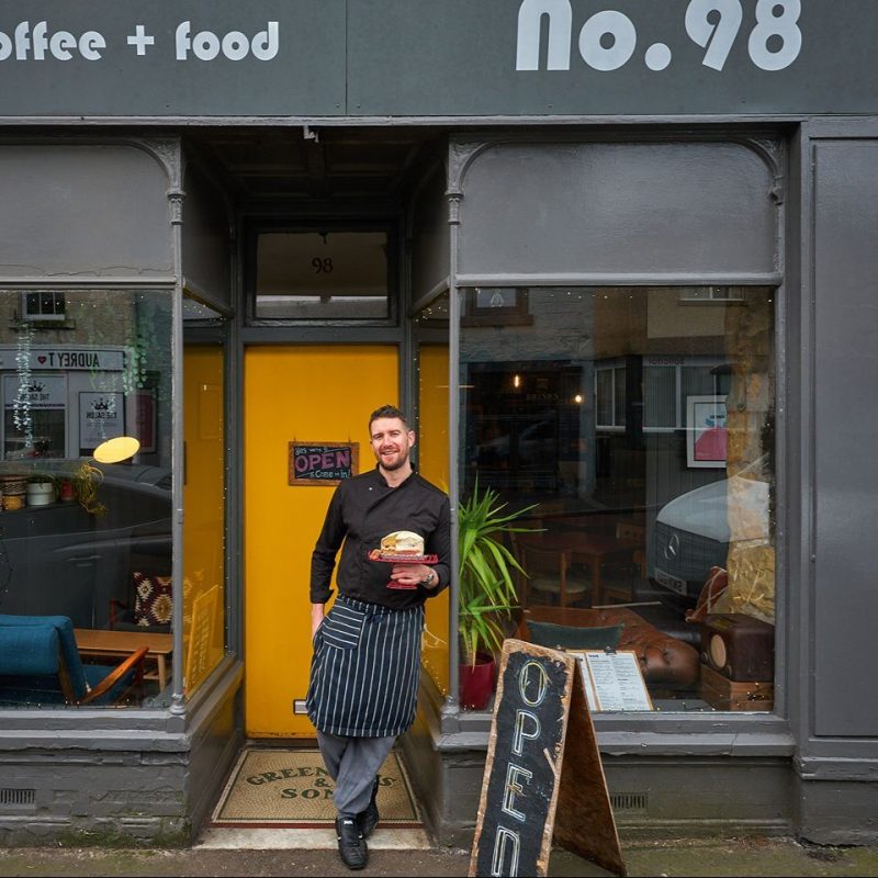 A smiling man wearing a shirt and apron holds a cake with a potion missing while leaning against a large window outside a cafe.