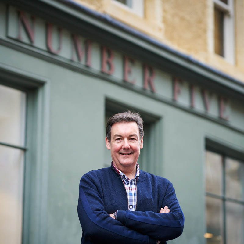 A smiling man wearing a blue jumper and checked shirt stands with his arms crossed outside a grey shop
