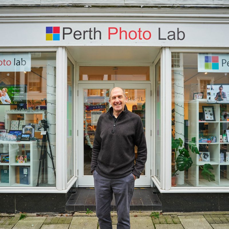 A smiling man wearing a black jumper stands outside a shop with two large windows displaying photography equipment in Perth