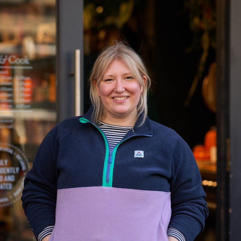 A smiling woman with blonde hair wearing a blue and pink jumper stands in a doorway outside a shop in Perth