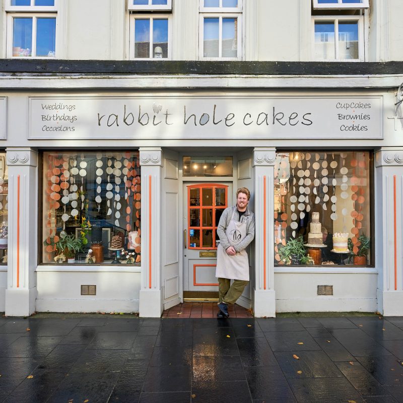 A smiling man wearing a grey apron leans against a grey and orange painted bakery with two windows displaying decorated cakes