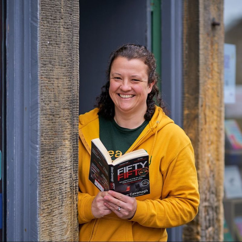 A smiling woman wearing a yellow jumper stands in a doorway and holds an open book