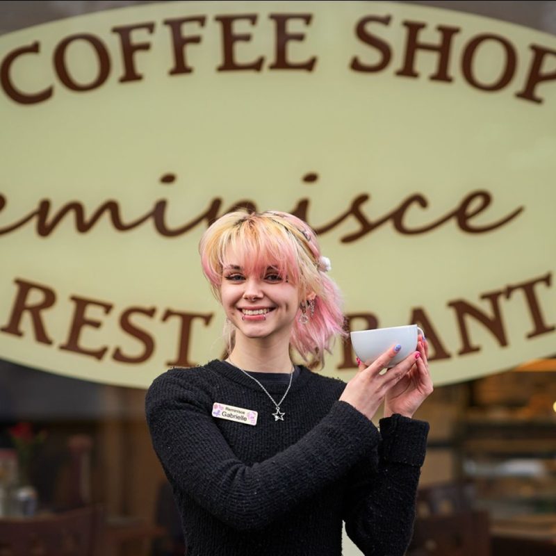 A smiling woman with pink hair wearing a black shirt holds a mug outside a large cafe window with a yellow and brown decal
