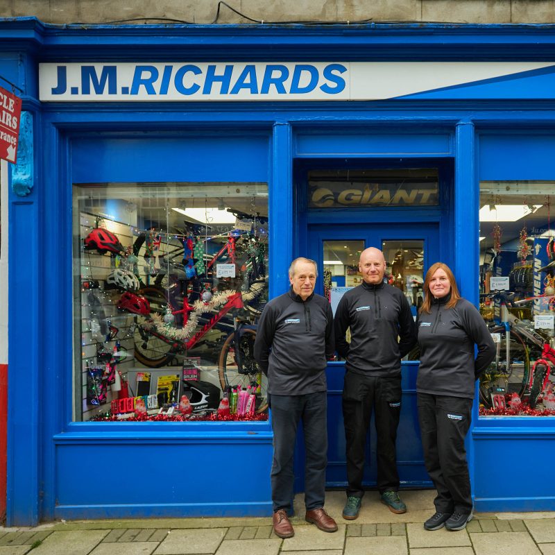 Two men and a woman wearing black stand together outside a blue shop with two windows displaying bikes and cycling supplies