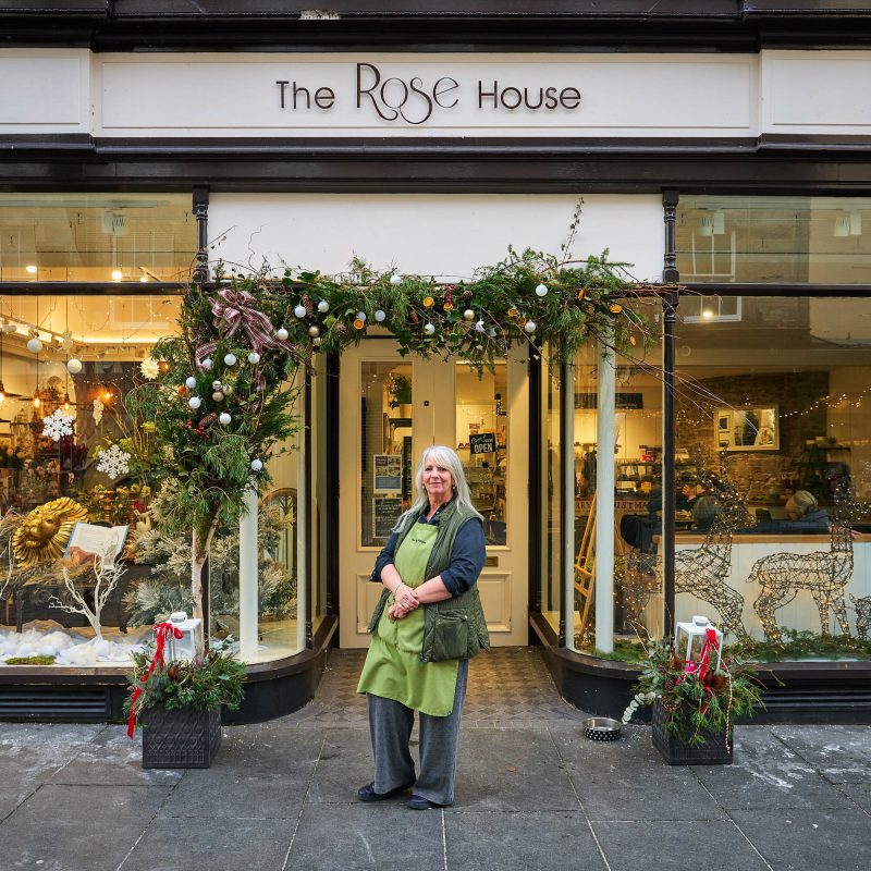 A woman wearing a green apron stands outside a cafe with two large windows decorated with baubles, ferns, and festive decorations