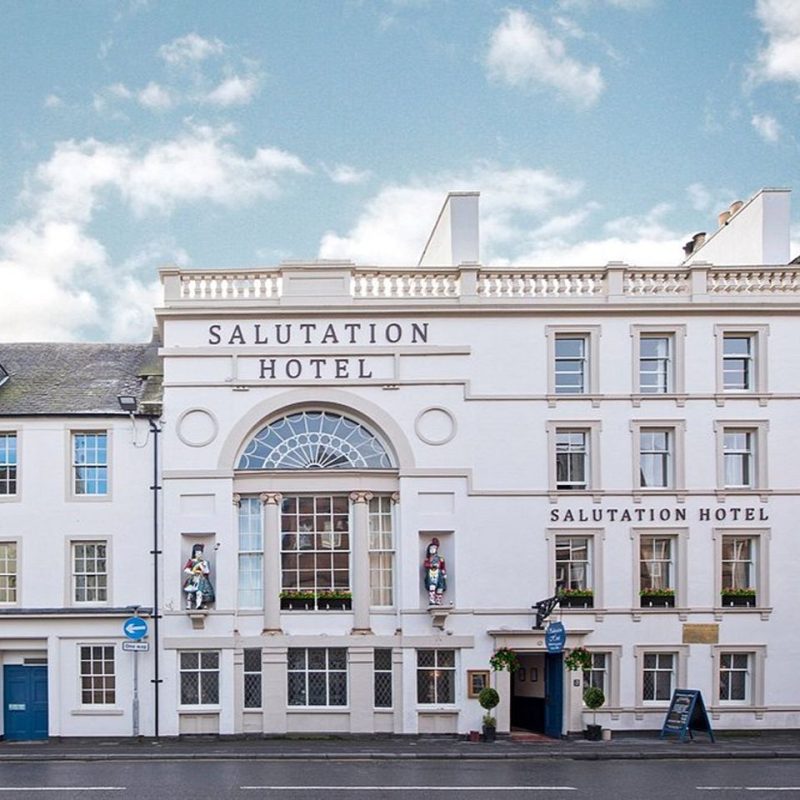A lavish white hotel with very large windows between two pillars with a statue of a Scottish Highland soldier on either side