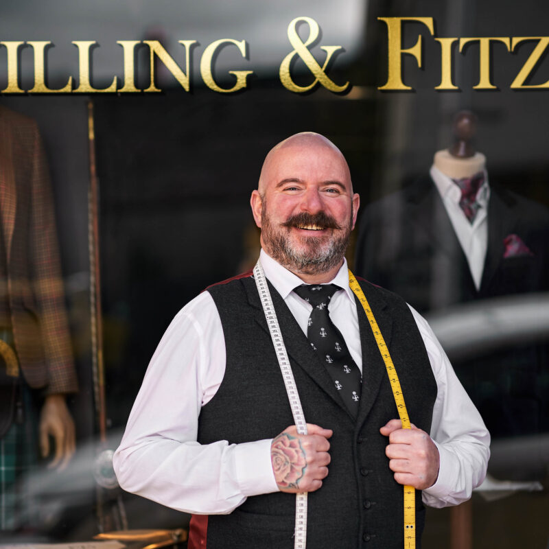 A smiling man with a dark moustache wearing a shirt and black drapes a measuring tape around his neck and stands outside a shop