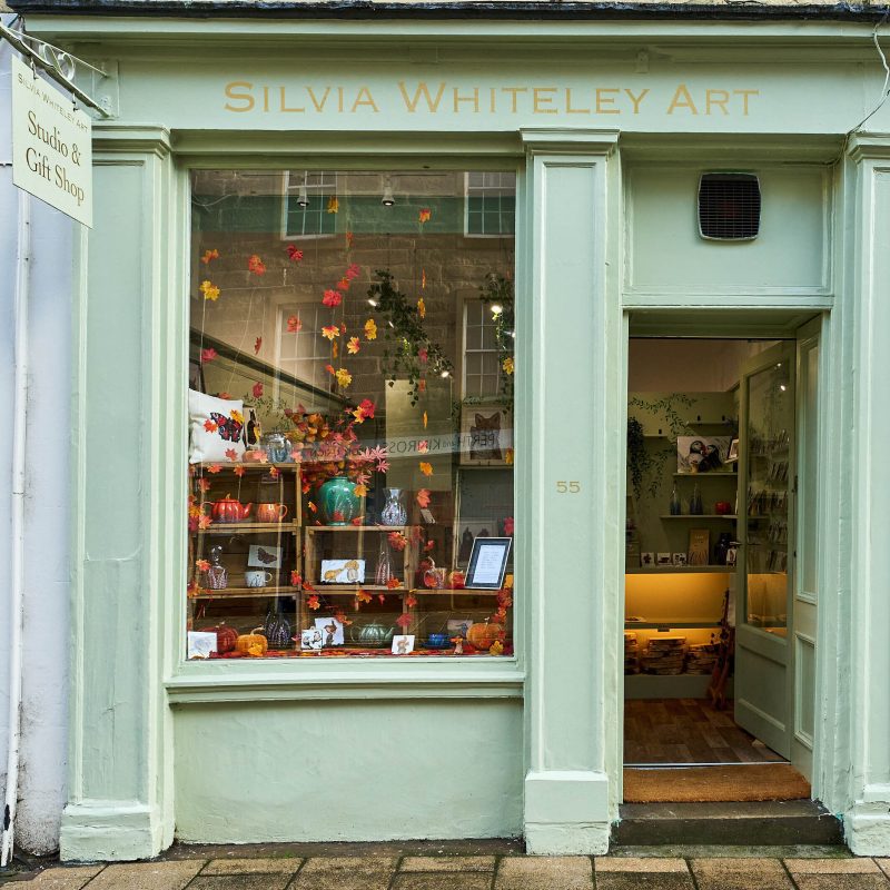 A pale green shop with gold lettering reading Silvia Whiteley Art above a large window decorated with autumnal leaves and art