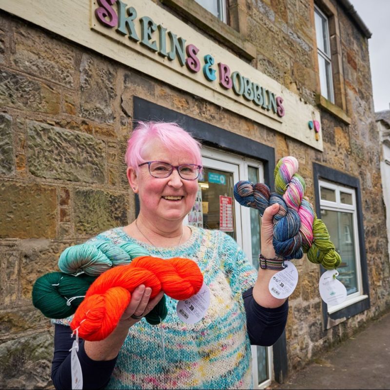 An older woman with pink hair wearing a blue jumper holds colourful yarn outside a shop with a multi-coloured sign