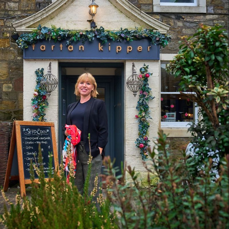 A smiling woman wearing a black shirt holds a brightly coloured scarf outside a shop with vines decorating the door and sign.