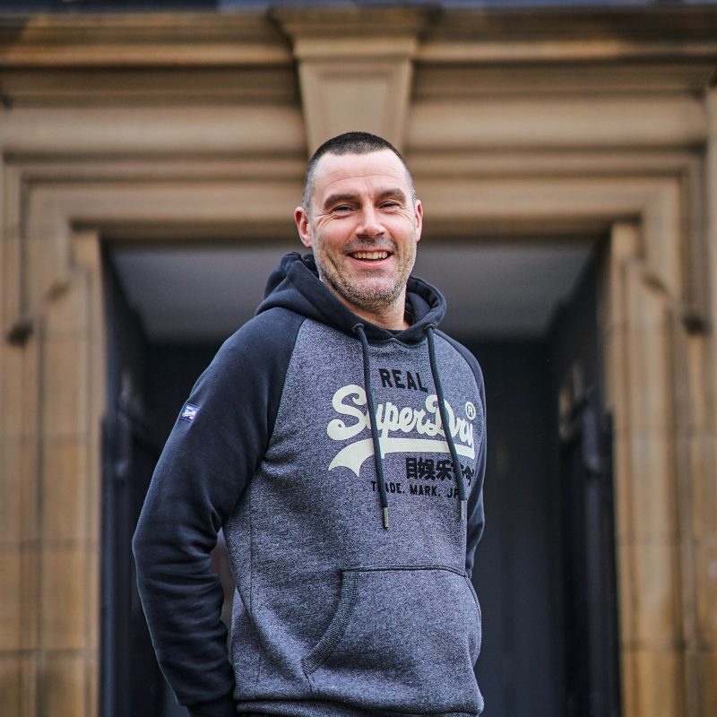 A smiling man wearing a grey jumper with his hands behind his back stands in front of a stone doorway