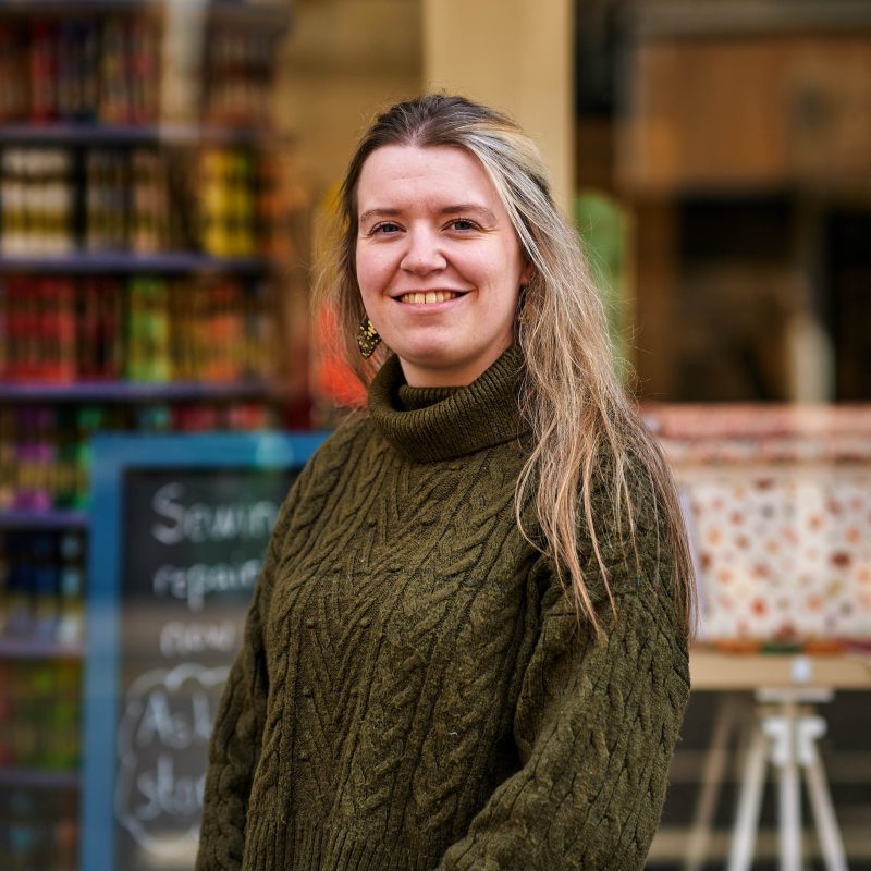A smiling woman with blonde hair wearing a green sweater stands outside a shop window with a blue sign