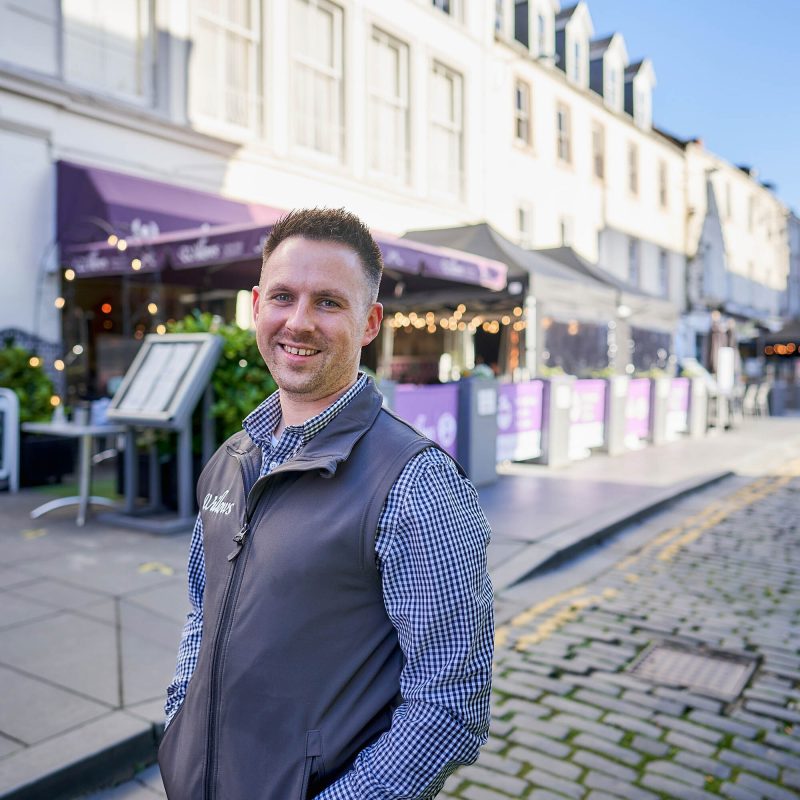 A smiling man wearing a grey shirt and jumper stands outside a cafe with purple signs and a large canopy in Perth