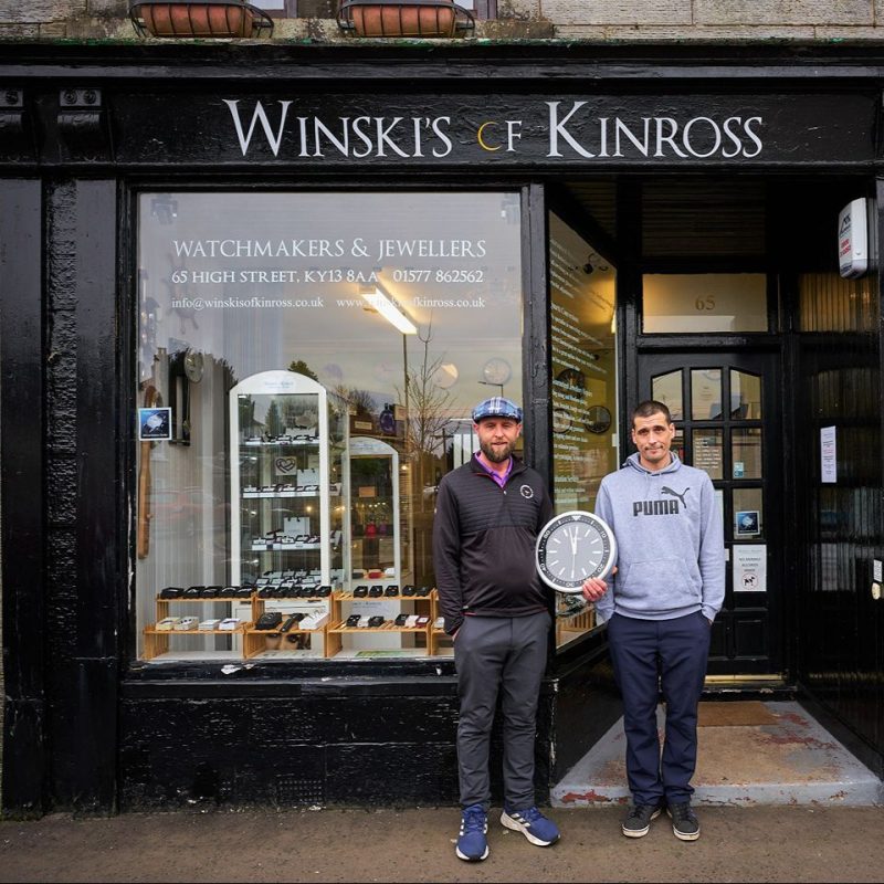 A man wearing a black shirt and holding a black clock stands next to a man wearing a grey shirt outside a black shop front