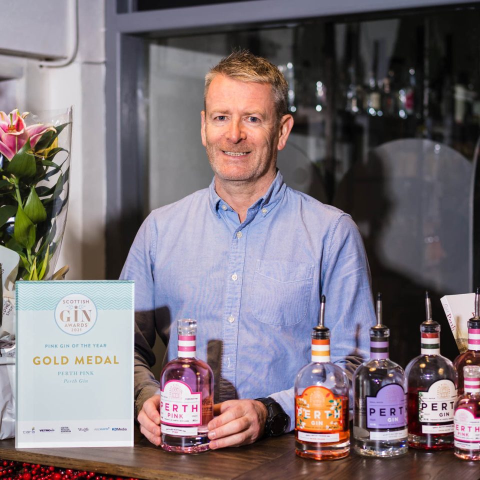 A smiling man wearing a blue shirt holds a bottle of pink gin and stands behind a counter displaying a sign and bottles of gin