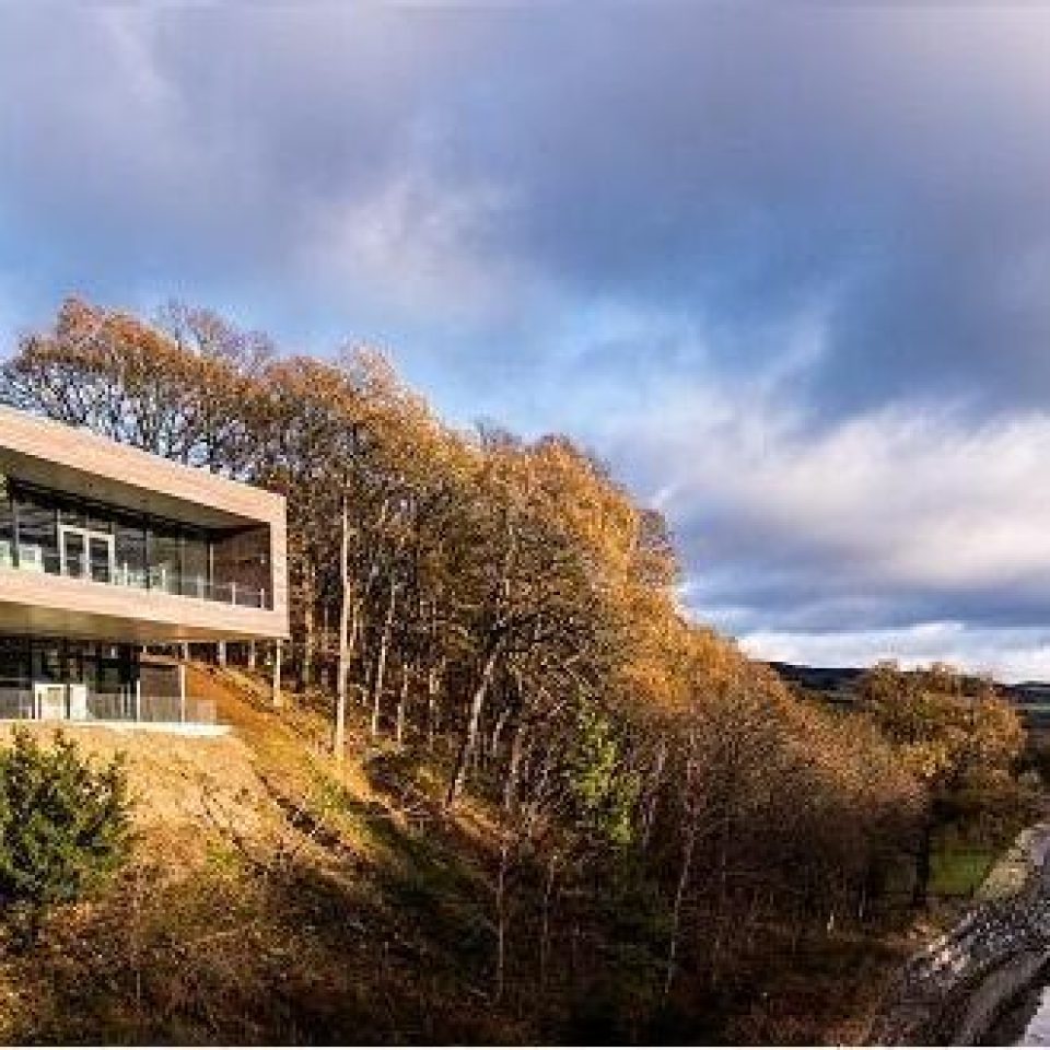A square building with a glass panelled balcony its among autumnal trees on a hill overlooking a river