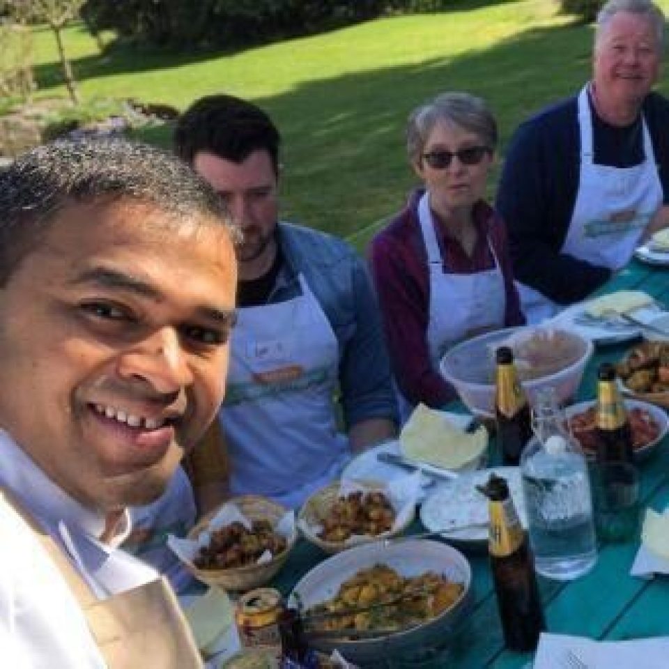 A group of people wearing matching white aprons sit around an outside bench covered in a spread of an array of food and wine